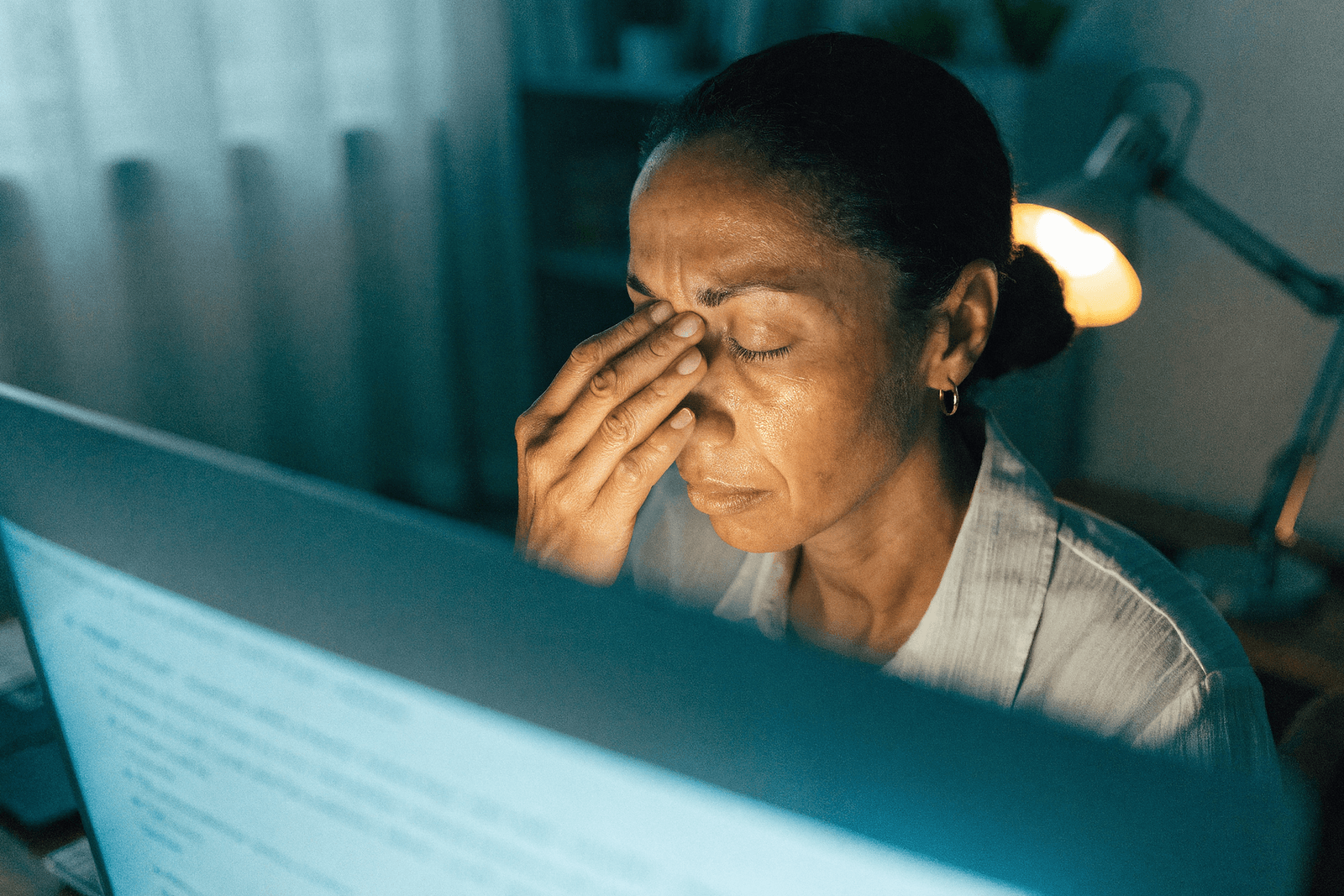 Mulher em frente ao computador à noite descansando os olhos cansados com a mão no rosto · ardor e olhos secos ligados ao uso de telas
